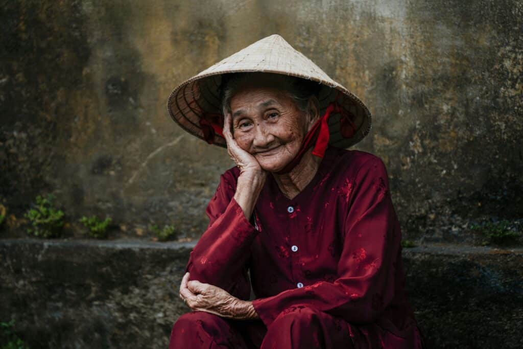 An elderly woman in a traditional conical hat and red outfit, smiling peacefully and sitting outdoors, symbolizing self-love, wisdom, and inner peace.