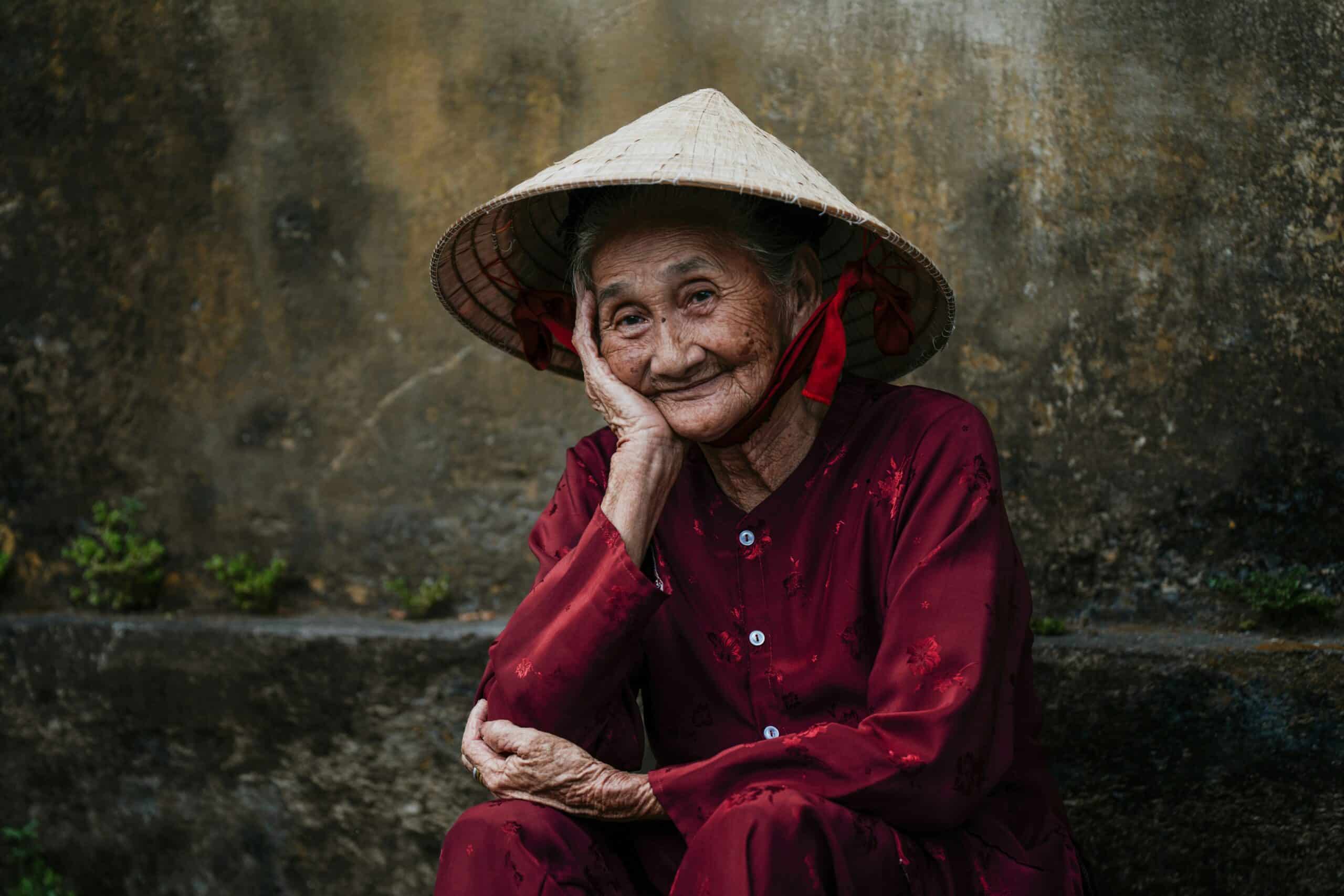 An elderly woman in a traditional conical hat and red outfit, smiling peacefully and sitting outdoors, symbolizing self-love, wisdom, and inner peace.