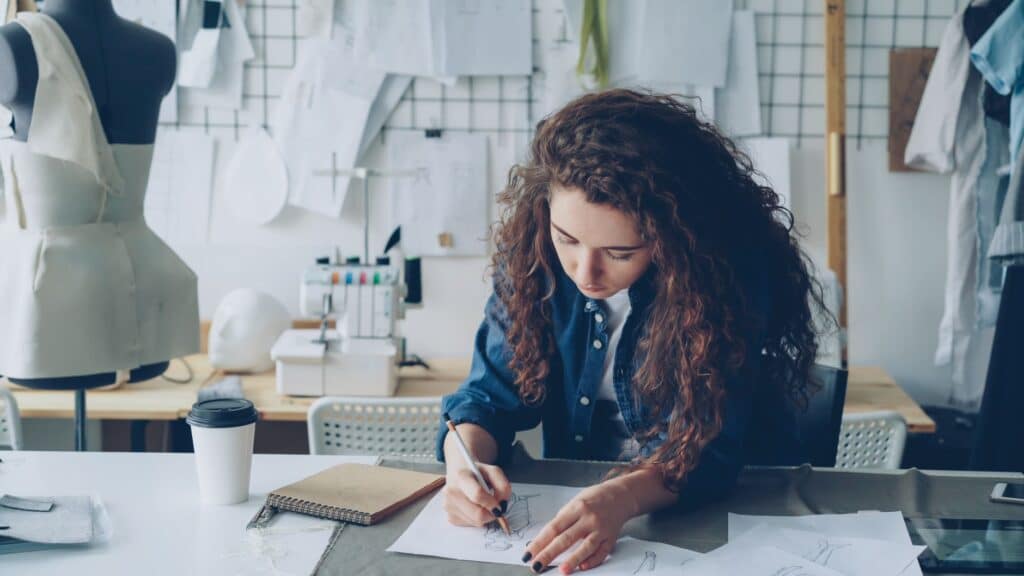 Young female fashion designer sketching in a stylish studio setting with mannequins and sewing equipment.